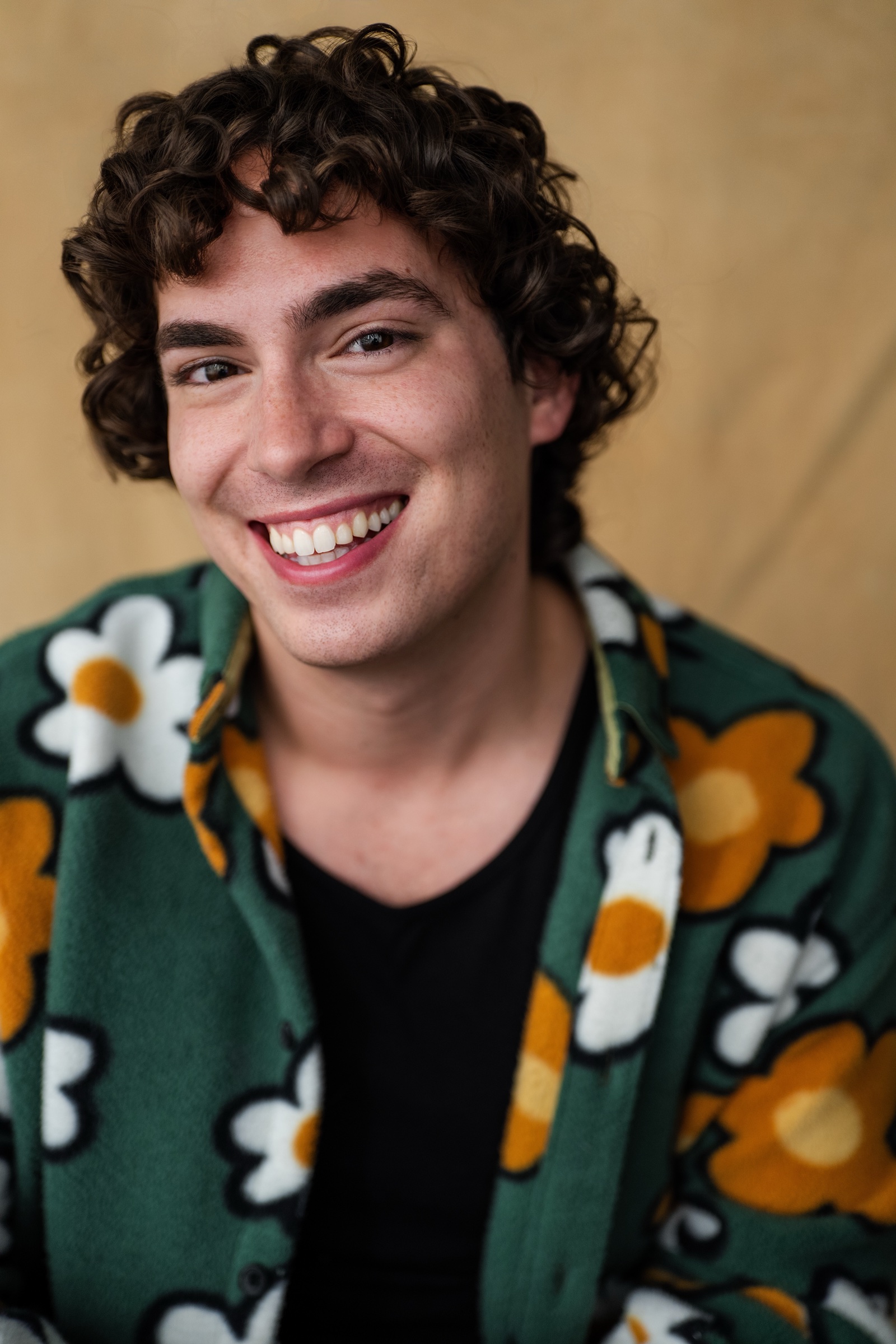 Neil Kelly headshot — laughing in a green floral button-down over a black tee against a warm beige backdrop