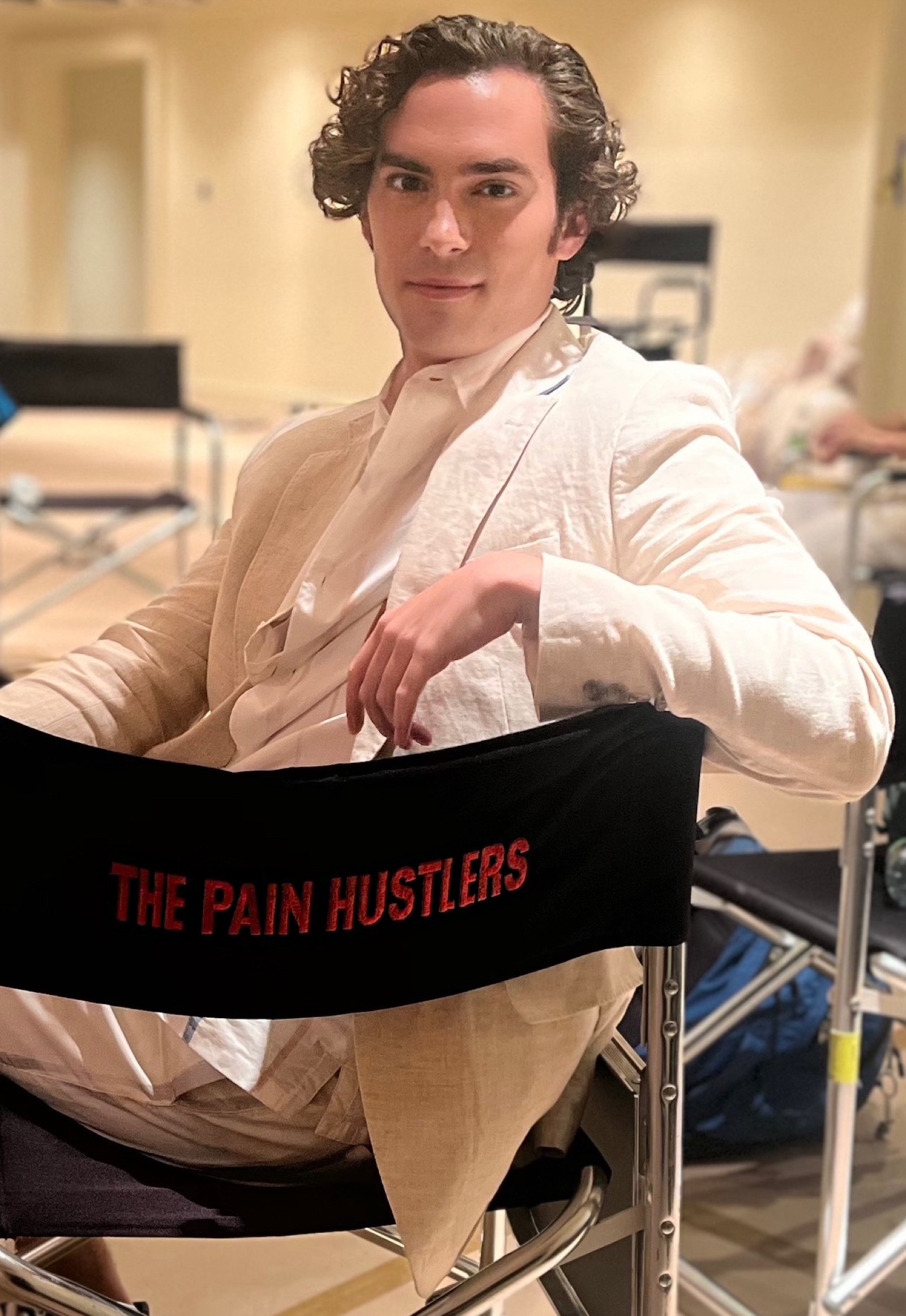 Neil Kelly on set of Pain Hustlers, seated in a director's chair with the production logo on the back