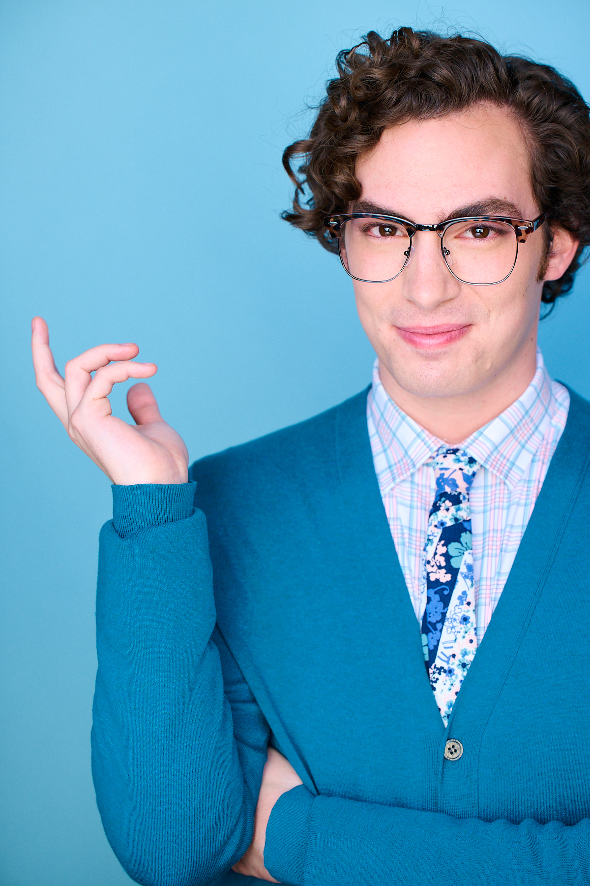Headshot of Neil in glasses and a teal cardigan over a floral tie, one hand raised, playful expression