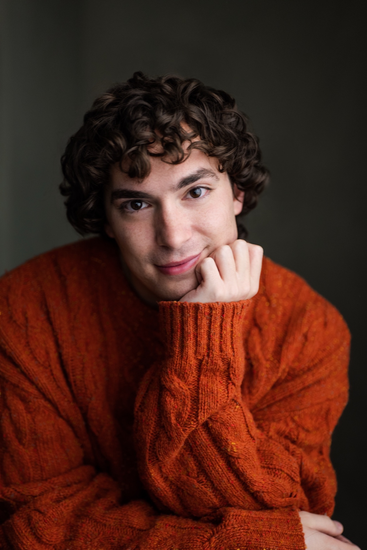 Neil, hand under chin, rust-orange cable-knit sweater on moody gray-green — warm thinker, soft direct gaze (portrait → crop landscape)