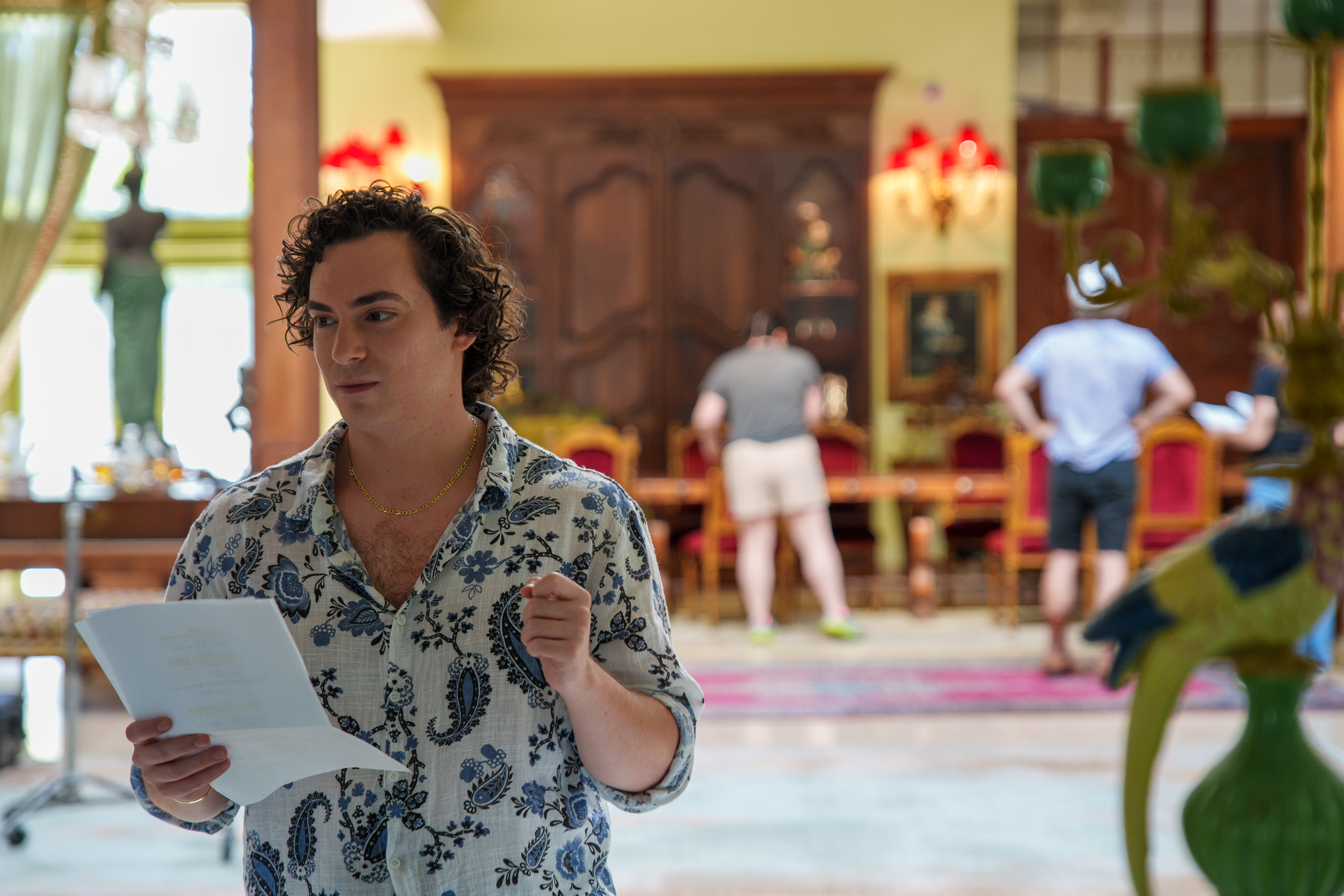 Neil on set, script pages in hand, glancing aside mid-thought while crew resets a table behind him