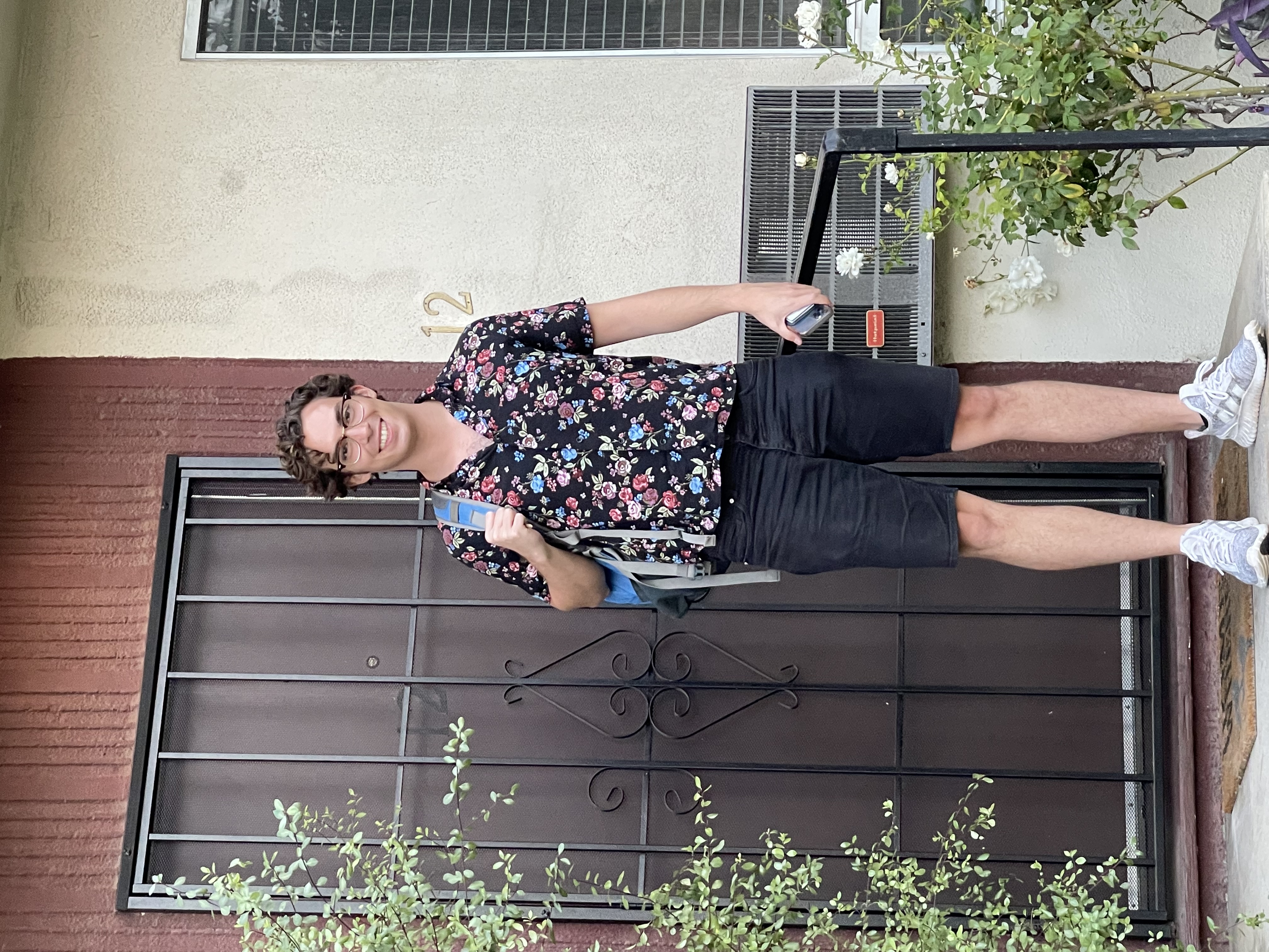 Neil in a floral shirt standing in front of an ornate black iron gate at a building entrance, smiling