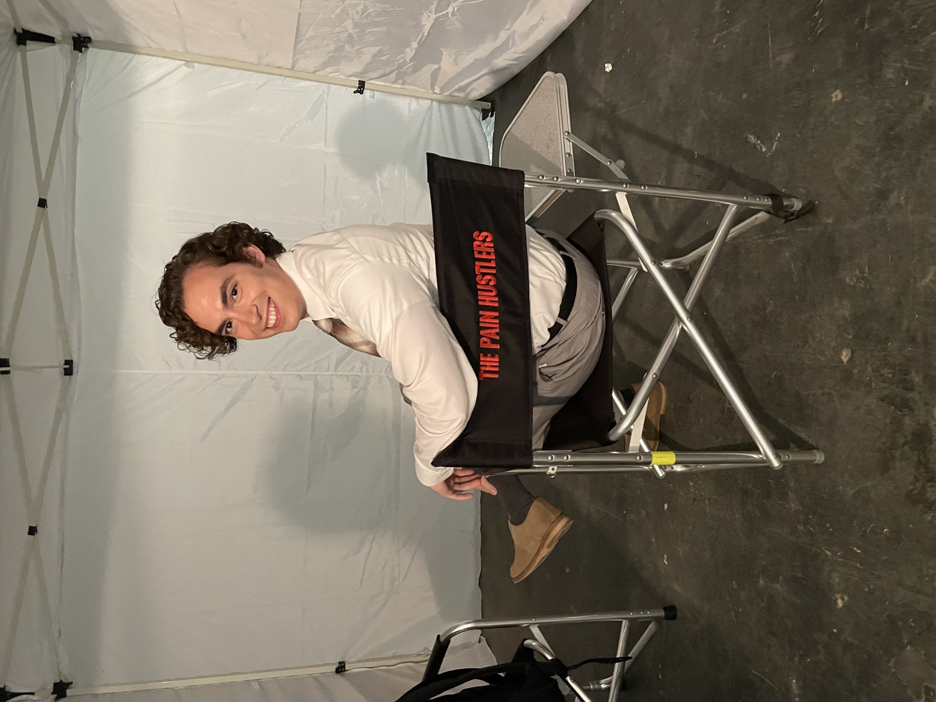 Neil in costume seated on a folding chair smiling over his shoulder; chair back reads "THE PAIN HUSTLERS" in red text
