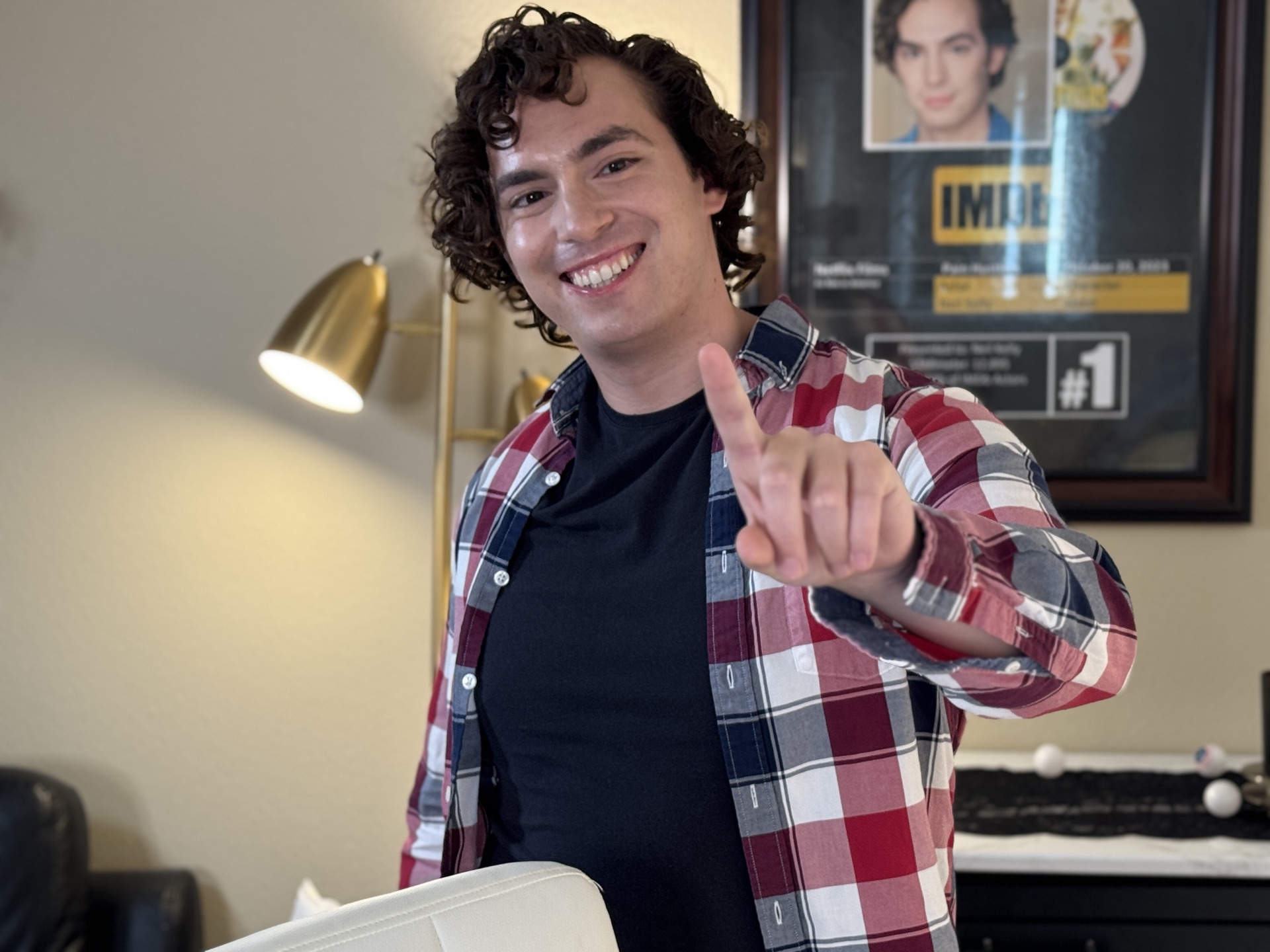 Neil in plaid flannel pointing at the camera with a wide grin, framed IMDb listing for Pain Hustlers (#1) on the wall behind him (landscape, face visible)